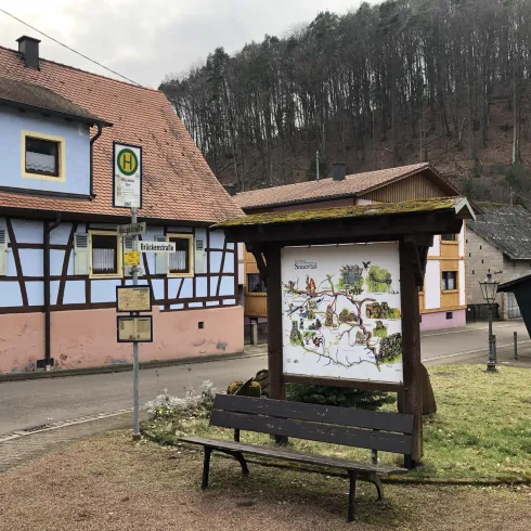 Eine Straße in einem kleinen Dorf mit farbenfrohen Häusern und einer Informationsstelle. Im Hintergrund sind bewaldete Hügel zu sehen.