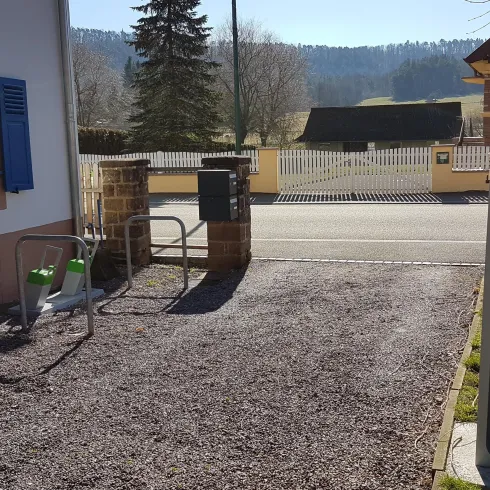 Ein Parkplatz mit einem Ladegerät für Elektroautos und einer Sitzgelegenheit. Im Hintergrund sind Bäume und ein ruhiger Weg zu sehen.