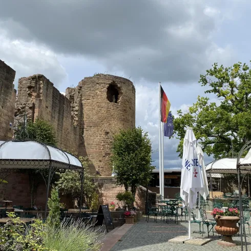 Eine alte Ruine mit einem Turm und einer deutschen Flagge. Im Vordergrund sind Tische und Stühle in einem Gartenbereich.