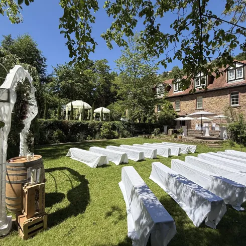 Ein schöner Garten mit weißen Tischen und Bänken unter einem blauen Himmel. Im Hintergrund sind Pavillons und ein Gebäude zu sehen.