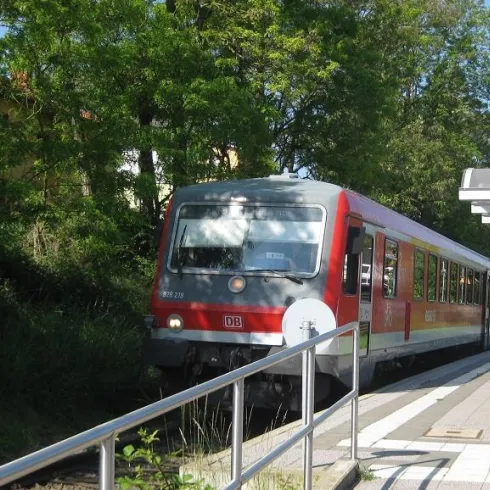 Ein roter Zug wartet an einer Haltestelle. Im Hintergrund sind Bäume und ein klarer Himmel zu sehen.