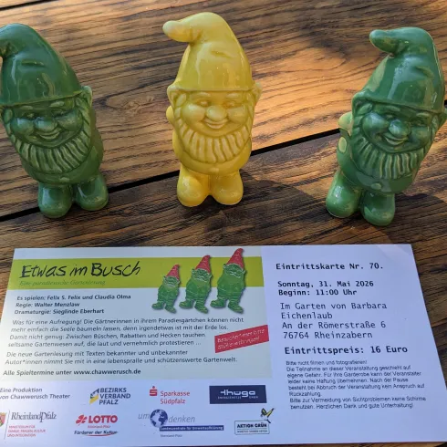 Drei bunte Gartenzwerge stehen auf einem Tisch. Daneben liegt eine Einladung zu einer Veranstaltung.