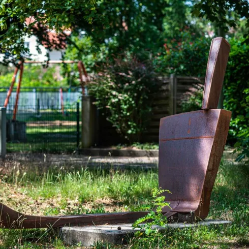 Eine rustikale Skulptur in Form eines Stuhls steht in einem Grünbereich. Umgeben von Bäumen und Gras wirkt die Szene ruhig und einladend.
