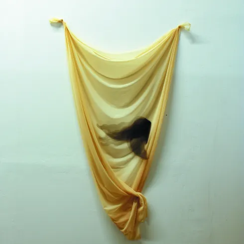Ein transparentes, gelbes Tuch hängt an der Wand und bildet sanfte Wellen. Dahinter ist eine schemenhafte Gestalt zu erkennen, die eine mysteriöse Atmosphäre schafft.