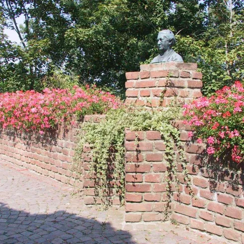 Eine Mauer aus Ziegelsteinen mit einem bekanntem Denkmal und bunten Blumen. Im Hintergrund sind Bäume und ein klarer Himmel sichtbar.