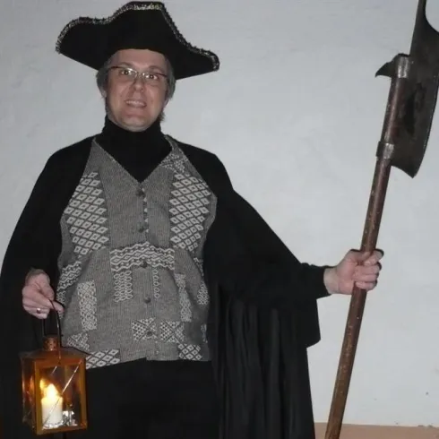 Eine Person in historischer Kleidung hält eine Laterne in der einen Hand und eine Axt in der anderen. Sie trägt einen großen Hut und steht vor einer neutralen Wand.