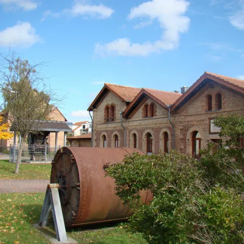 Ein historisches Gebäude mit roten Ziegelfassaden und einem nahegelegenen großen Rad. Der Himmel ist blau mit einigen Wolken, umgeben von Bäumen und Rasen.