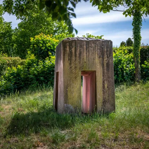 Ein rechteckiger Steinskulptur inmitten von grünem Gras und Büschen. Die Skulptur hat Öffnungen in der Mitte und wird von Bäumen umgeben.