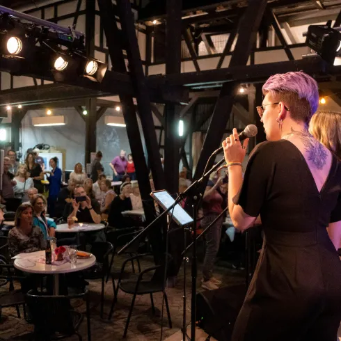 Eine Sängerin steht auf der Bühne und singt in einem lebhaften Veranstaltungsraum. Das Publikum sitzt an Tischen und ist aufmerksam.