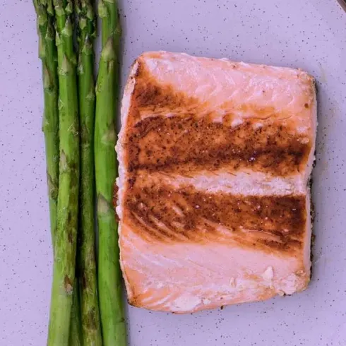 Ein Stück gebratener Lachs liegt auf einem weißen Teller. Daneben befinden sich frische grüne Spargelstangen.
