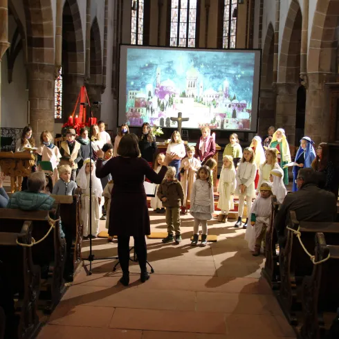 Eine Aufführung in einer Kirche mit vielen Kindern, die in bunten Kostümen stehen. Im Hintergrund ist eine große Leinwand mit einer bunten Szene zu sehen.