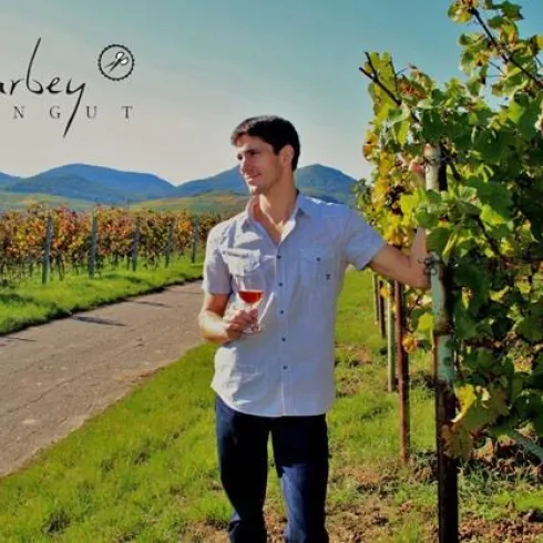 Ein Mann steht in einem Weinberg und hält ein Glas Roséwein in der Hand. Im Hintergrund sind grüne Reben und Hügel zu sehen.