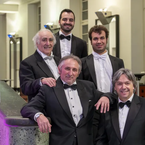 Fünf Männer in eleganter Abendgarderobe stehen lächelnd zusammen. Die Szene wirkt festlich und gehoben.