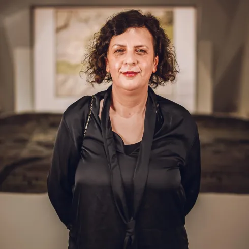 Eine Frau steht lächelnd in einem Ausstellungsraum. Im Hintergrund sind Gemälde oder Kunstwerke zu sehen.