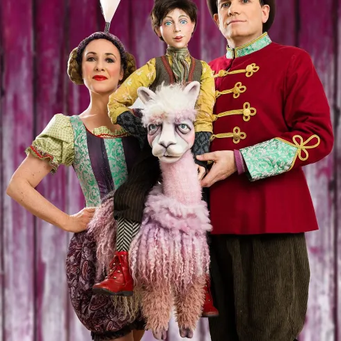 Eine bunte Kunstszene mit zwei Darstellern in Zirkus Kostümen. Sie stehen vor einem rosa Hintergrund und der Mittelpunkt ist ein verkleidetes Lama.