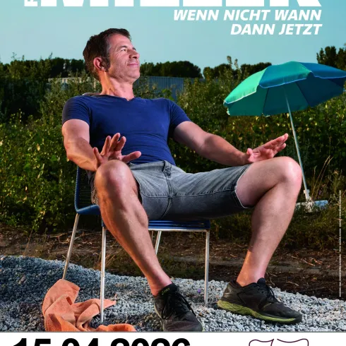 Ein Mann sitzt entspannt auf einem Stuhl im Freien und genießt die Sonne. Im Hintergrund sind grüne Felder und ein Sonnenschirm zu sehen.