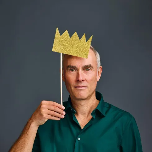 Ein Mann steht vor einem grauen Hintergrund und hält einen goldenen Kronen-Stick in der Hand. Er trägt ein grünes Hemd und hat einen neutralen Gesichtsausdruck.