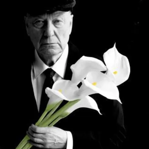 Ein älterer Mann trägt einen schwarzen Anzug und einen Hut. In seiner Hand hält er weiß-blühende Lilien vor einem dunklen Hintergrund.