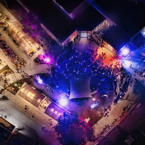 Eine lebendige Veranstaltung bei Nacht mit bunten Lichtern und vielen Menschen. Stände und ein großes Zelt sind im Zentrum des Geschehens zu sehen.