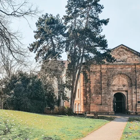 Ein historisches Gebäude mit einer beeindruckenden Fassade befindet sich in einer grünen Umgebung. Hohe Bäume rahmen den Eingang und verleihen der Szenerie eine ruhige Atmosphäre.