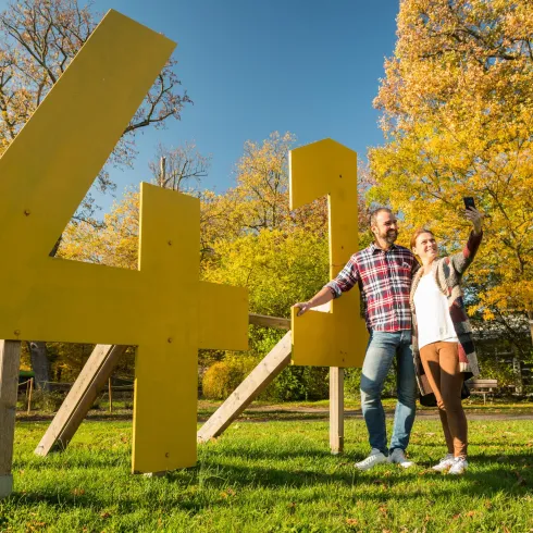 Ein junges Paar macht ein Selfie vor großen, gelben Zahlen im Freien. Im Hintergrund sind herbstliche Bäume zu sehen.