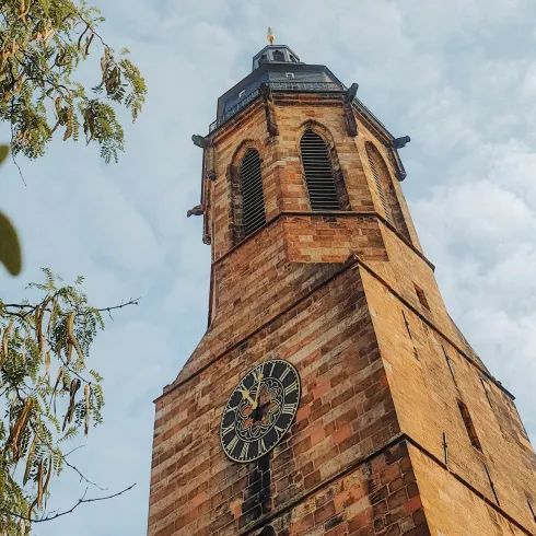 Ein hoher Kirchturm aus rotem Stein mit einer großen Uhr. Der Himmel ist bewölkt und es gibt grüne Blätter im Vordergrund.