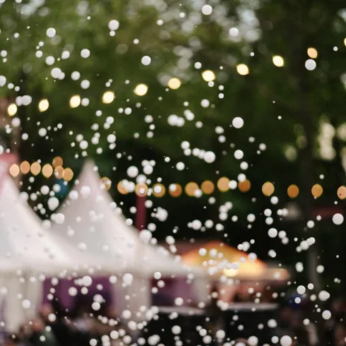 Ein festliches Ambiente mit glitzernden Lichtern und Zelten. Im Vordergrund sind viele helle Punkte zu sehen, die eine festliche Stimmung erzeugen.