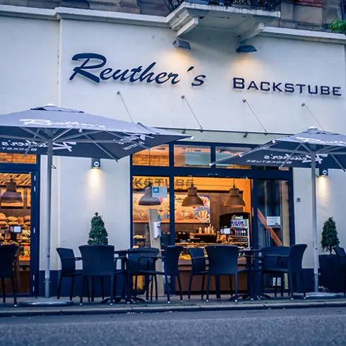 Ein einladendes Café mit dem Namen "Reuther's Backstube". Vor dem Gebäude stehen Tische und Stühle unter großen Sonnenschirmen.