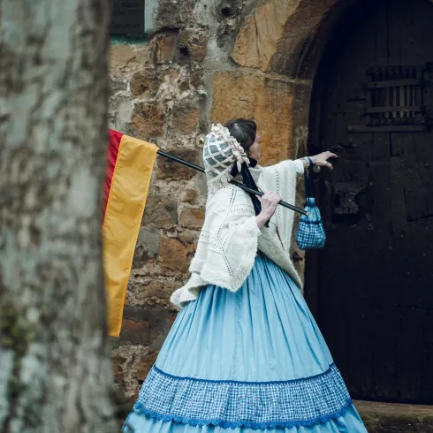 Eine Person in einem historischen Kleid steht vor einer alten Tür. Sie trägt eine Laterne und zeigt in eine Richtung.