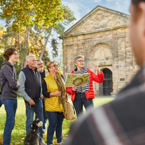 Eine Gruppe von Personen steht im Freien und hört einer Führerin zu, die einen Plan in der Hand hält. Im Hintergrund ist ein historisches Gebäude und Bäume im Herbst zu sehen.