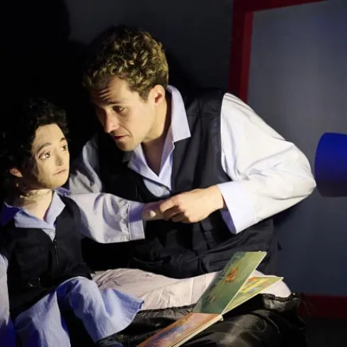 Ein Mann sitzt mit einer Puppe und liest ihr ein Buch vor. Die Szene ist in schwaches Licht getaucht, was eine intime Atmosphäre schafft.