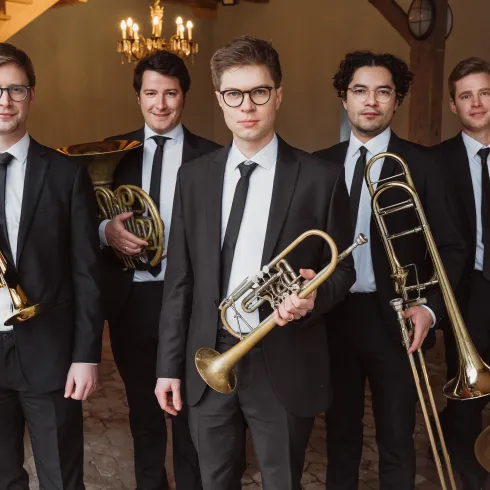 Eine Gruppe von fünf Musikern in Anzügen hält verschiedene Blasinstrumente. Sie stehen aufrecht vor einer stilvollen Kulisse mit einer eleganten Atmosphäre.