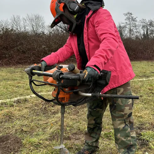 Eine Person mit Schutzkleidung bedient eine Erdbohrer auf einem offenen Feld. Der Himmel ist bewölkt und die Umgebung ist grün mit einigen Hecken im Hintergrund.