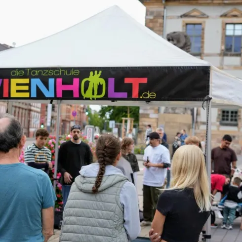 Eine Tanzschule präsentiert ihre Aktivitäten unter einem Zelt auf dem Platz. Menschen versammeln sich, um zuzuschauen und daran teilzunehmen.