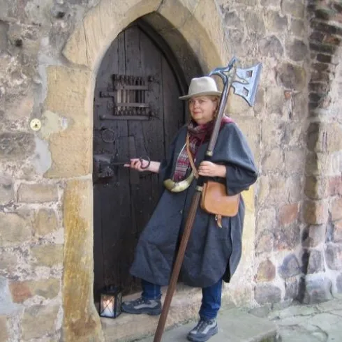 Eine Person steht vor einer alten, steinernen Tür und trägt eine historische Kleidung mit einem Hut und einer Axt. Im Hintergrund ist eine rustikale Mauer aus Steinen sichtbar.