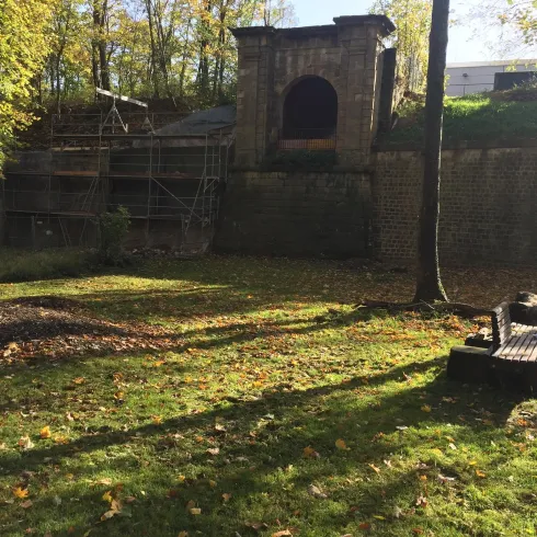 Eine alte Ruine im Wald mit herbstlichen Bäumen im Hintergrund. Die Umgebung ist grün und sonnig, mit Schatten auf dem Boden.