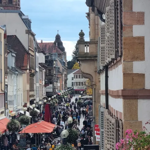 Eine belebte Straße mit vielen Menschen und Tischen im Freien. Im Hintergrund sind historische Gebäude und ein klarer Himmel zu sehen.