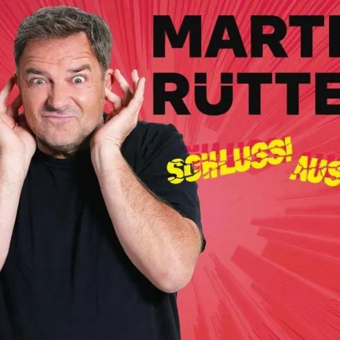 Ein Mann hält sich die Ohren zu und sieht überrascht aus. Im Hintergrund sind zwei bellende Hunde und der Titel "Martin Rütter Live" zu sehen.