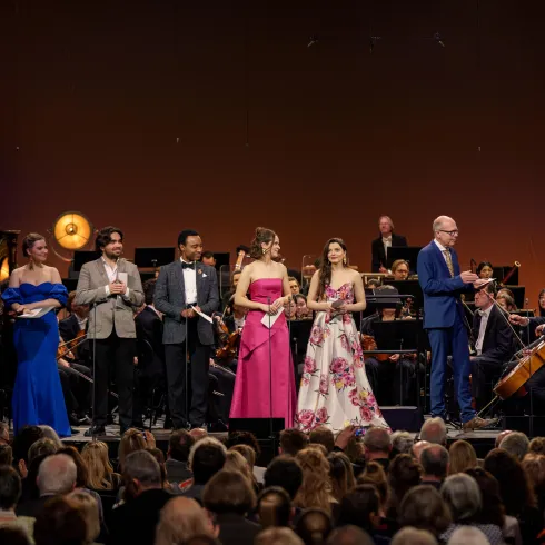 Eine Gruppe von Sängern und Moderatoren steht auf der Bühne vor einem Orchester. Das Publikum sitzt im Vordergrund und schaut gespannt zu.