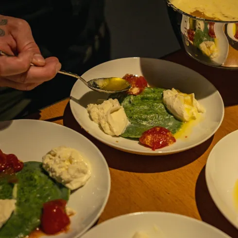 Ein Teller mit grünem Püree, Sahne und Tomatensoße. Eine hand, die mit einem Löffel eine Portion auf den Teller gibt.