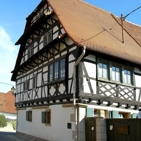 Ein traditionelles Fachwerkhaus mit einem roten Satteldach und großen Fenstern. Das Gebäude steht an einer ruhigen Straße und hat einen grünen Zaun.