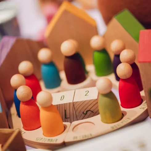 Bunte Holzfiguren stehen in einem Kreis auf einer Holzplatte. Im Hintergrund sind andere Spielzeuge sichtbar.