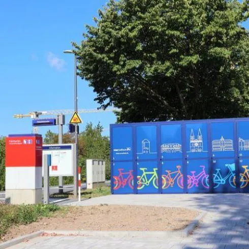 Eine Fahrradstation mit bunten Fahrradmotiven. Im Hintergrund befinden sich Bäume und ein klarer Himmel.