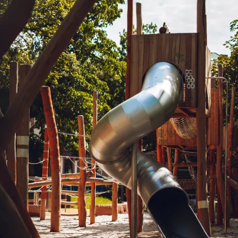 Ein moderner Spielplatz mit einer silbernen Rutsche und Holzelementen. Bäume umgeben den Spielbereich und bieten Schatten.