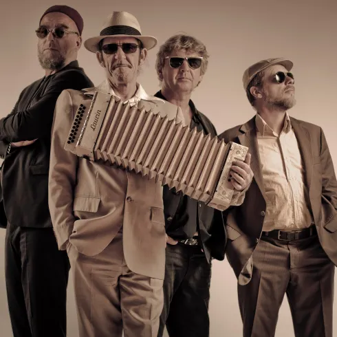 Vier Frauen in eleganten Kleidern sitzen um ein Klavier. Sie lächeln und posieren zusammen für das Foto.