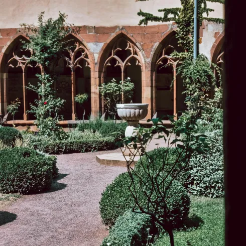 Ein ruhiger Garten mit gepflegten Buchsbäumen und grünen Pflanzen. Im Hintergrund sind gotische Fenster mit Steinbögen sichtbar.