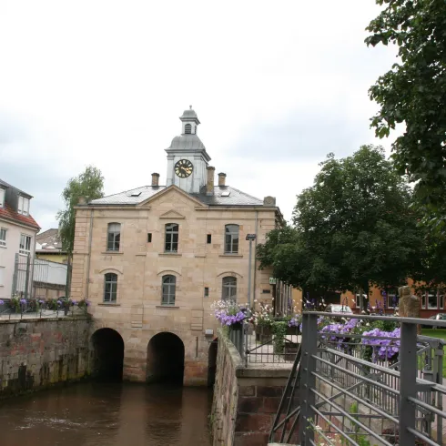 Ein historisches Gebäude mit einer Uhr an der Fassade, das über einen kleinen Kanal gebaut ist. Auf der linken Seite sind grüne Bäume und Blumen zu sehen.