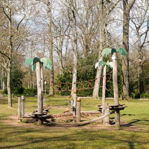 Ein Spielplatz mit Klettergerüst und palmenartigen Elementen. Im Hintergrund sind große, kahle Bäume und eine grüne Wiese sichtbar.