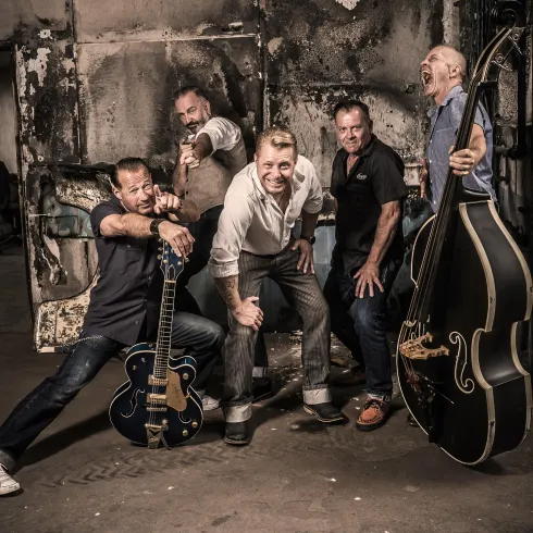 Eine Gruppe von fünf Männern steht in einem alten, industriellen Raum. Sie posieren spielerisch mit Musikinstrumenten und haben Spaß.