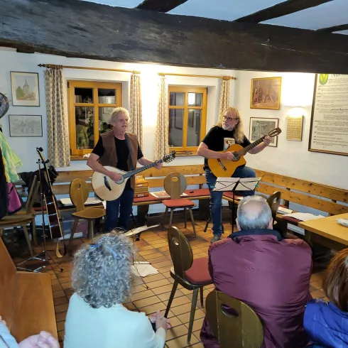 Zwei Musiker spielen Gitarre in einem gemütlichen Raum. Eine kleine Menge von Zuhörern hört aufmerksam zu.